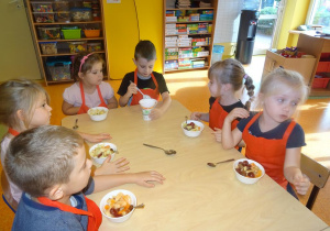 Pięcioro dzieci siedzi przy stole z wypełnionymi owocami miseczkami. Chłopiec nakłada jogurt.